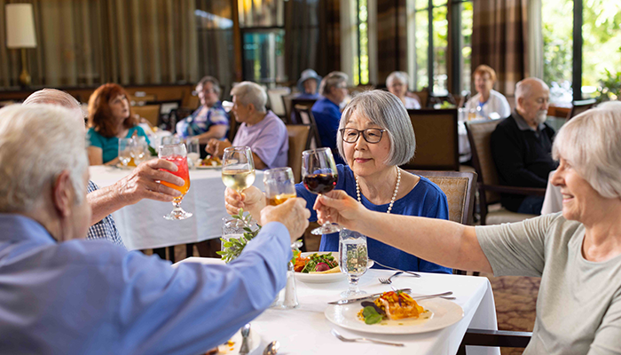 A group of older adults cheers glasses over dinner in a full dining room.