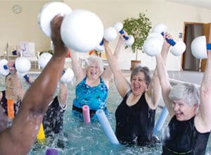 A group of older adults hold up water weights while floating on pool noodles.