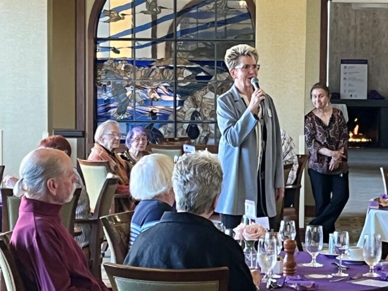 Woman in blue coat speaks in microphone to people seated at tables