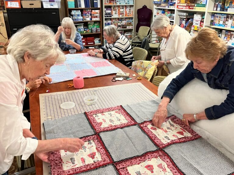 Women gather around a table sewing on small quilts