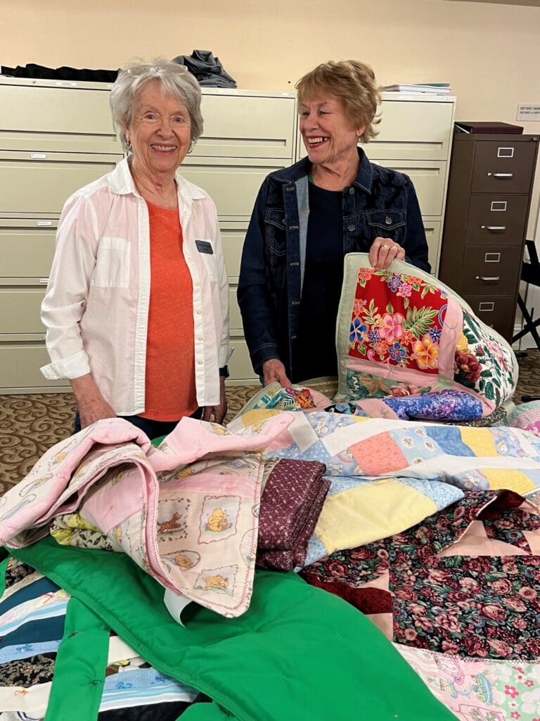 Two older women stand smiling behind a pile of hand-made quilts