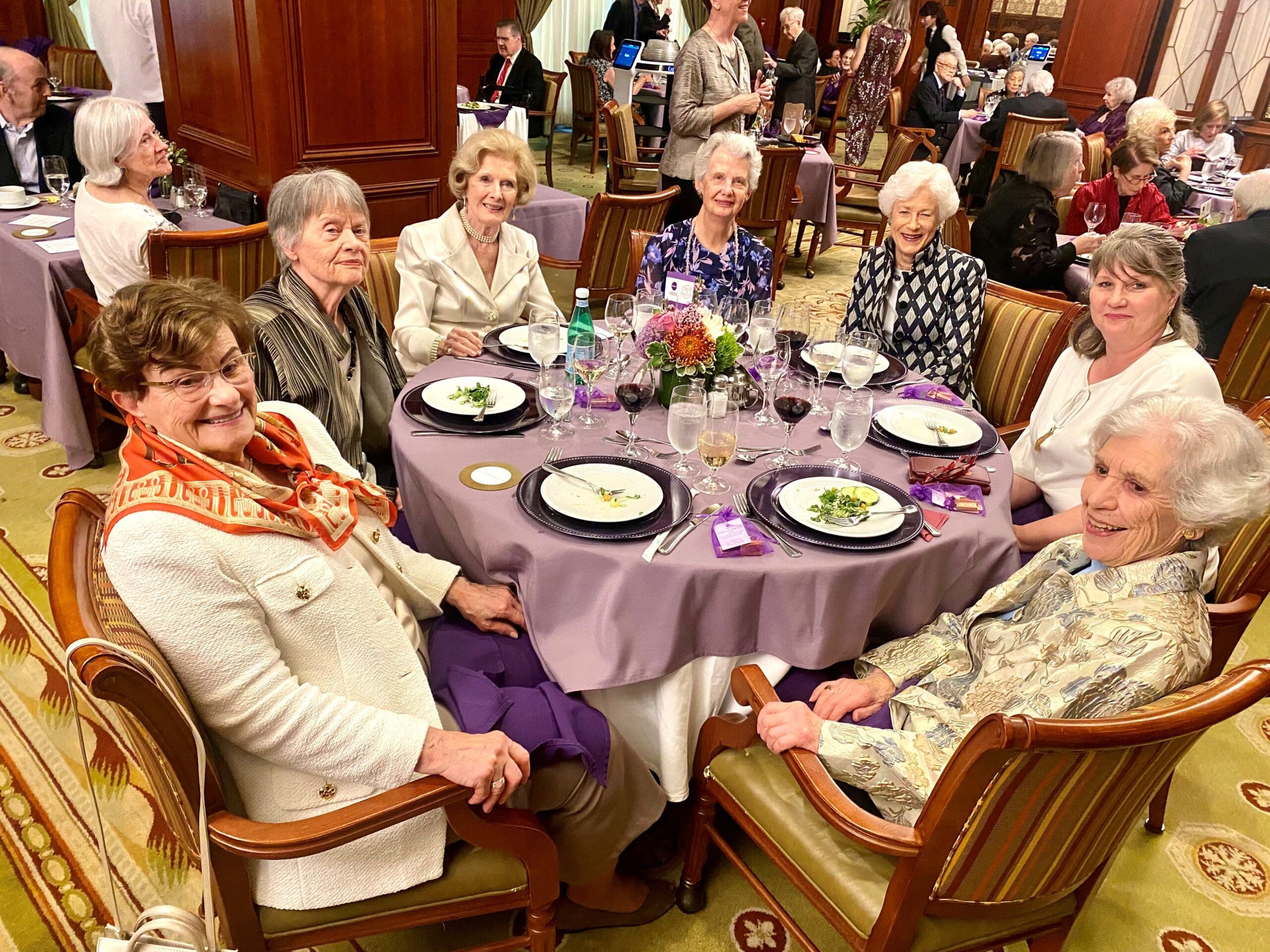 A group of women sit around a round dining table set with a lavender table cloth, plates, and glasses.