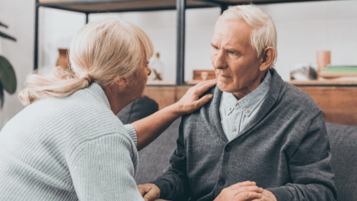 Two older adults look at each other. One sits on a couch and the other crouches in front of them.