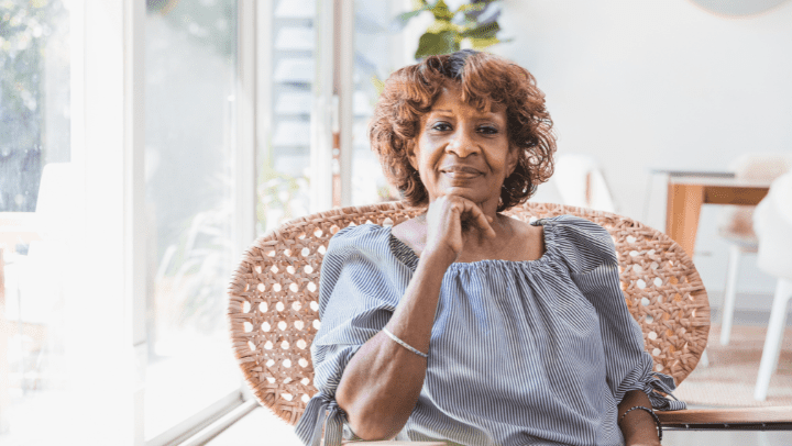 An older woman sits in a chair next to bright windows.