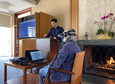 An older woman sits in a lounge wearing a VR headset. A person stands at a podium to her right.