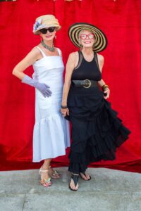 Two women pose in dresses and hats in front of a red backdrop