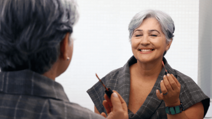 An older woman smiles in a mirror while applying make-up.