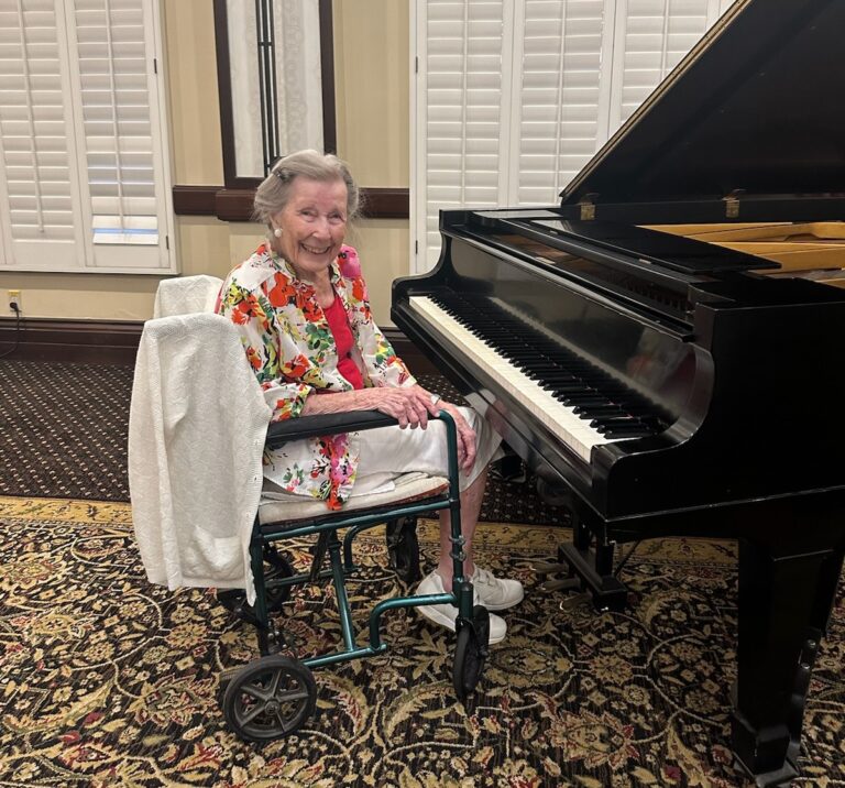 An older woman sits at a piano while smiling at the camera.