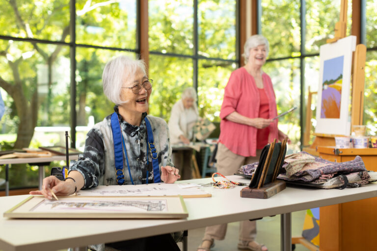 A group of people working on art projects in an art studio with big windows that showcase green trees and natural light