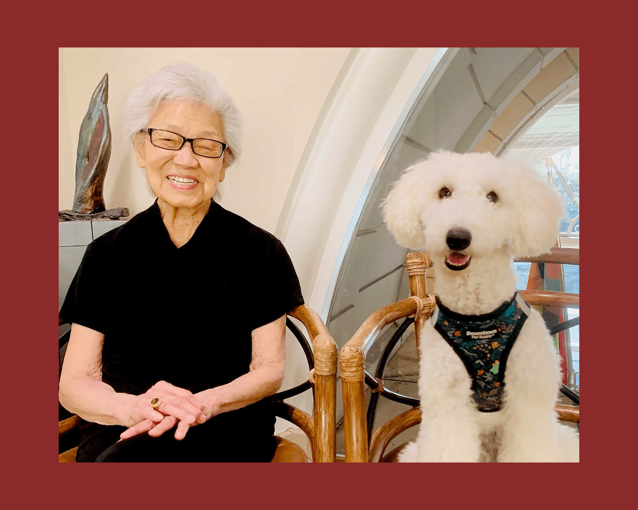 An older woman sits in a chair next to a white fluffy dog also sitting in a chair. Both smile for the camera.