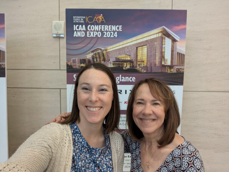 Two women stand in front of a poster for the ICAA Conference and Expo 2024