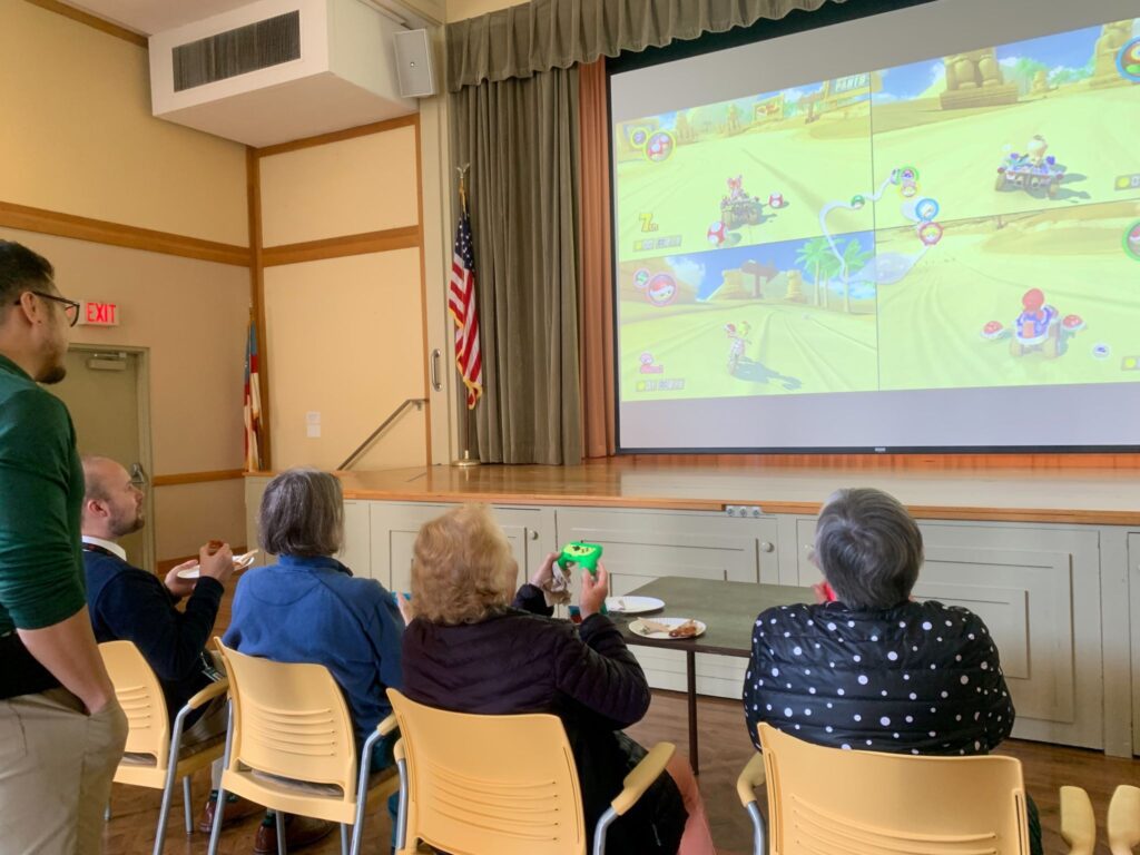A group of older adults play a video game. They look up at a large screen on a stage. Two staff members watch as they play, sitting beside and standing behind them.