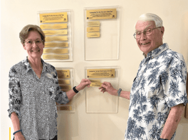 Two older adults hold up a placard in front of a wall set with placards.