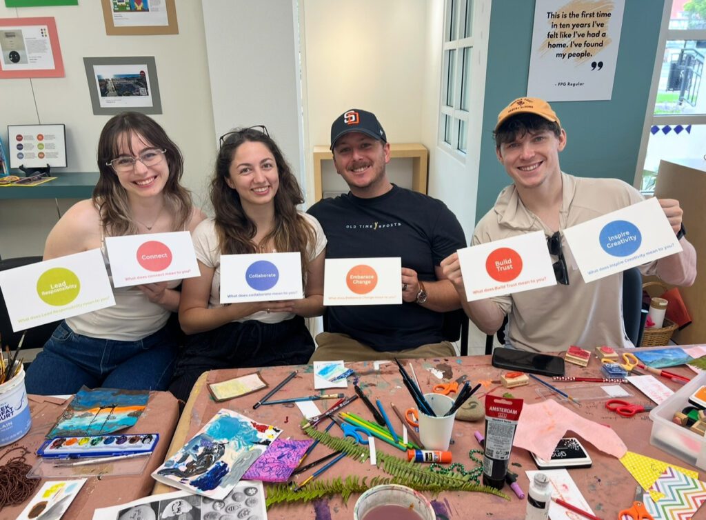 A group of people sit at a table set with art supplies. Each holds a standee featuring a colorful circle.