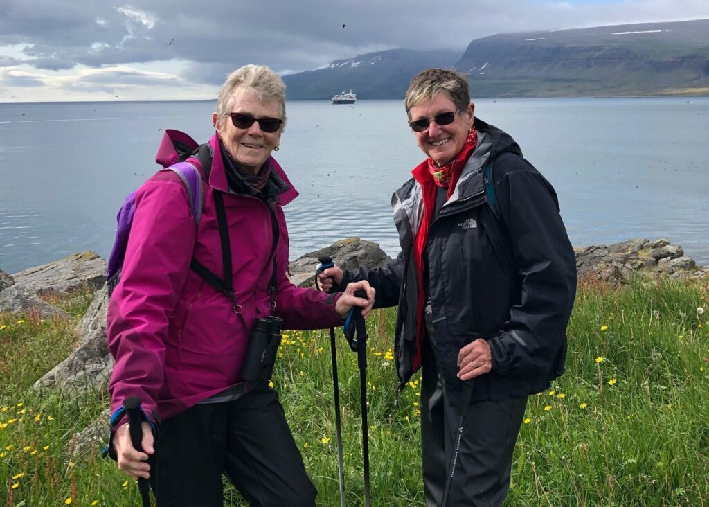 Two older adults pose on a grassy cliff with views of the water behind them.