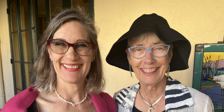 Two women wearing glasses smile together. One wears a black hat.