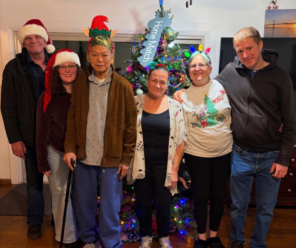 A group of people wearing winter holiday accessories pose in front of a Christmas tree. A blue key that says "Home Match" hangs from the top of the tree.