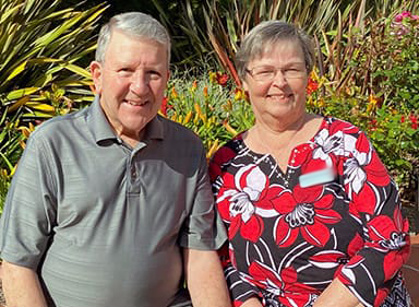 Two older adults sit outside in front of plants.
