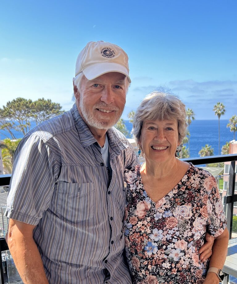 Two older adults stand in front of a view of the ocean.