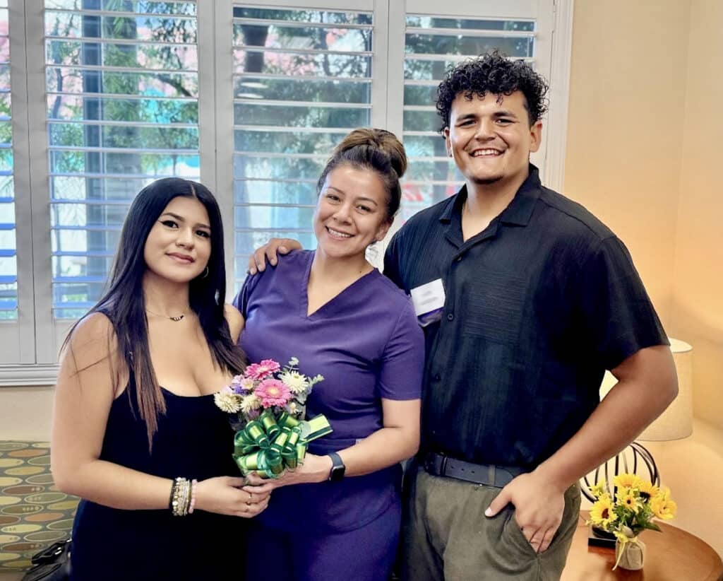 Three People Pose Together. One Person Holds A Small Bouquet Of Flowers.