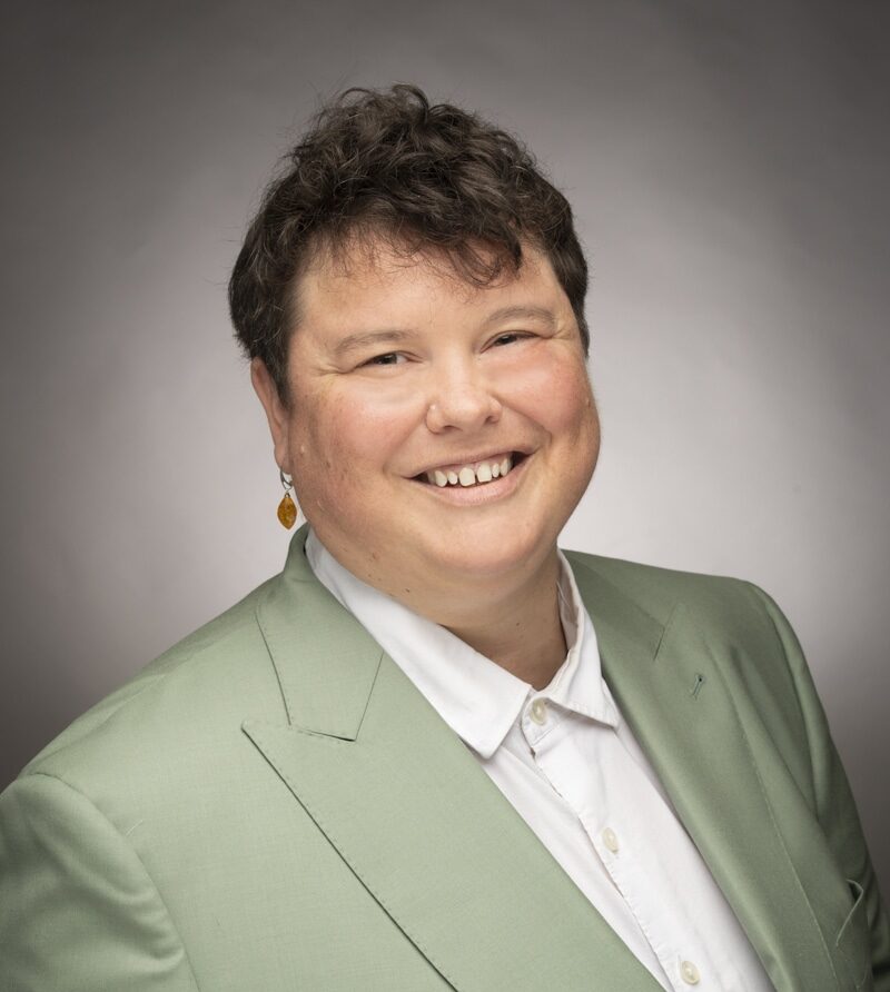 A Person With Short Hair Wearing A White Shirt And Pale Green Blazer.