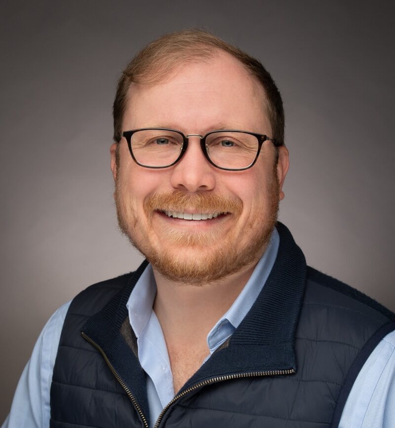 A Person With Glasses Wearing A Blue Shirt And Vest.