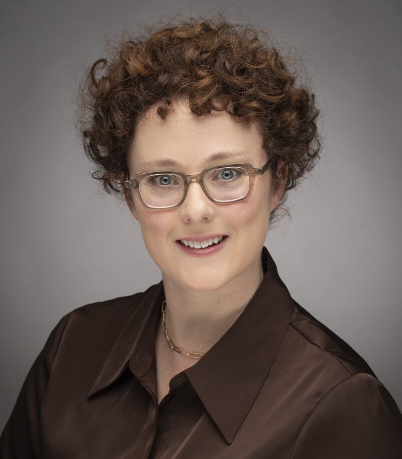 A Person With Short Curly Hair And Glasses, Wearing A Brown Button-Up Shirt.