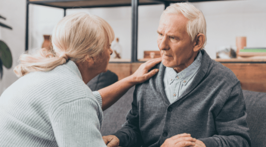 Two older adults look at each other. One sits on a couch and the other crouches in front of them.