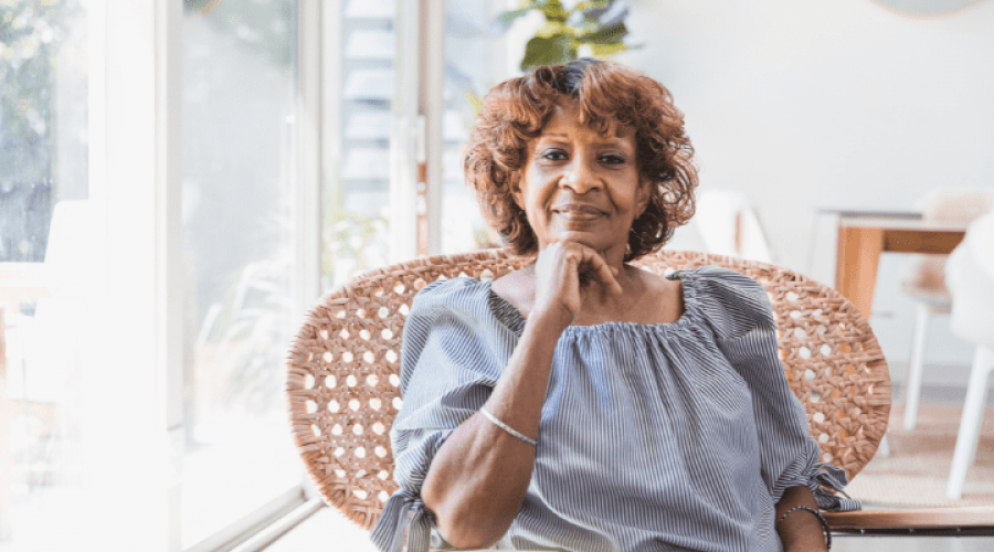 An older woman sits in a chair next to bright windows.