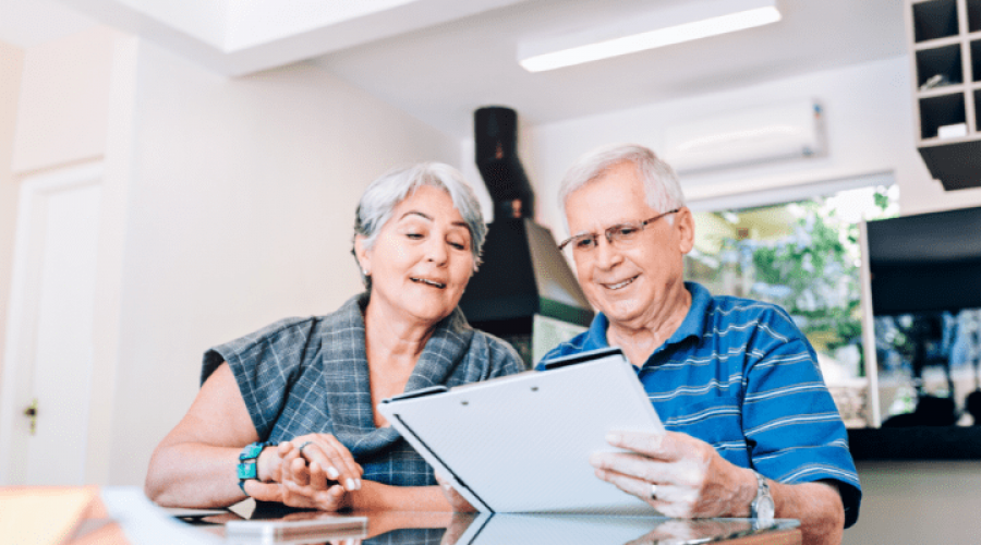 Two older adults sit at a table in a kitchen, looking at paperwork.