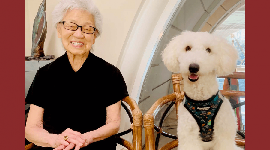 An older woman sits in a chair next to a white fluffy dog also sitting in a chair. Both smile for the camera.