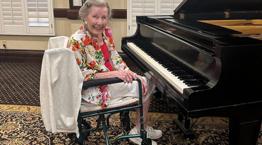 An older woman sits at a piano while smiling at the camera.