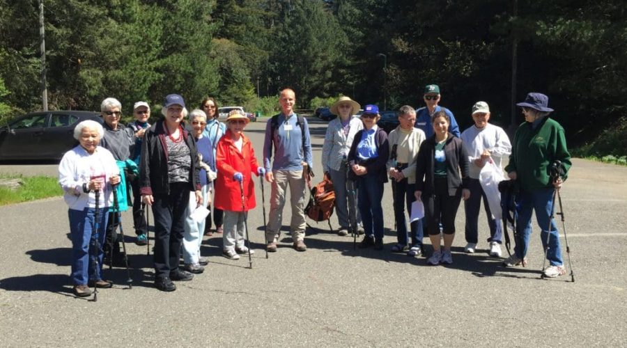 A group of older adults pose outside in a parking lot with a forest in the background.