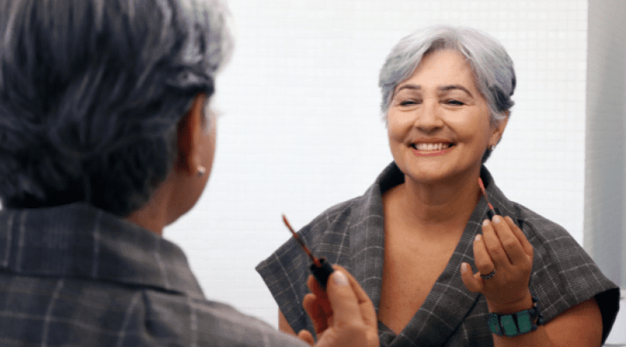 An older woman smiles in a mirror while applying make-up.