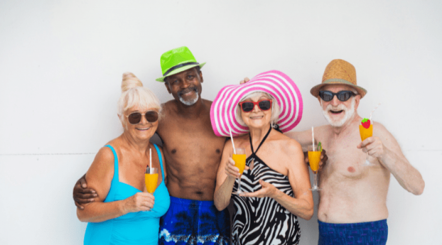 A group of older adults wearing bathing suits smile while holding up orange drinks.